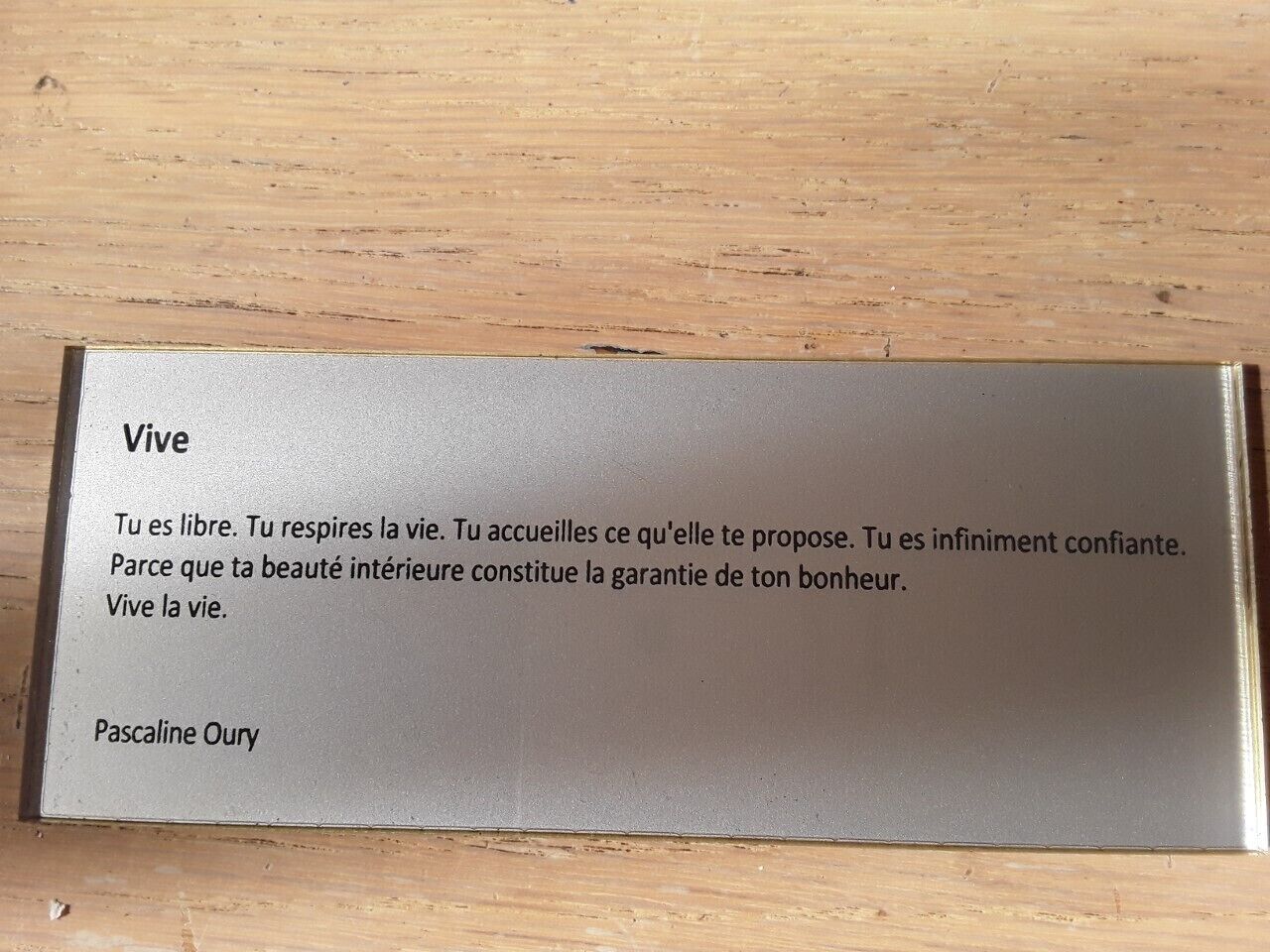 Een houten oppervlak met een metalen plaquette met een citaat van Pascaline Oury.