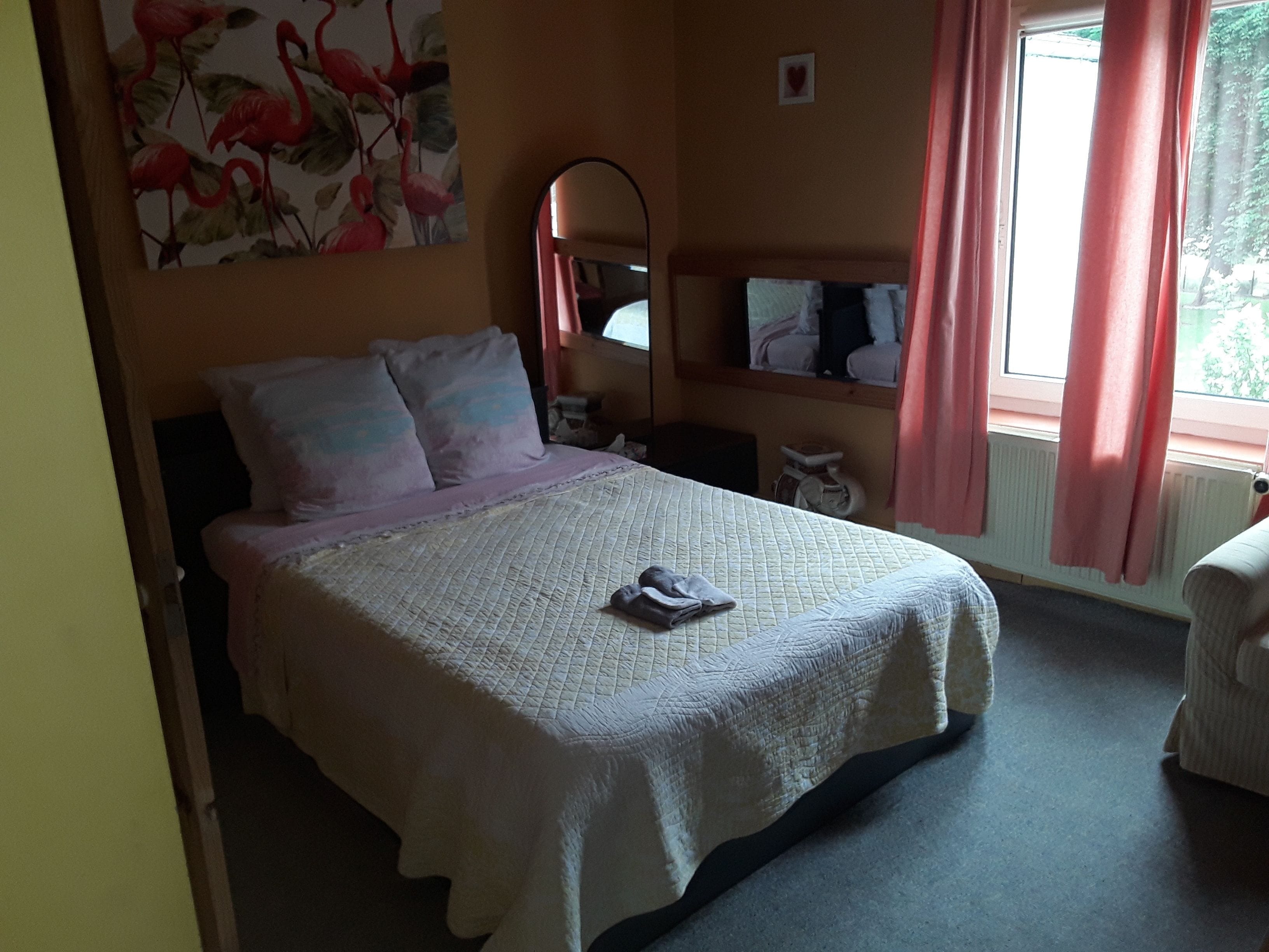 A bedroom with a bed, pillows, a bedside table, a mirror, and a window with curtains.
