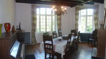 A dining area with a piano, wooden chairs, a table with a tablecloth, and large windows with patterned curtains.