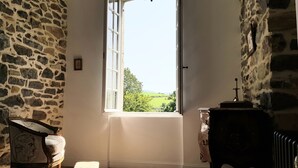 A room with stone walls, a window with a view of greenery, a radiator, and a chair with a decorative cushion.