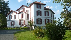 A large, two-story building with red shutters and a white facade, surrounded by lush greenery and a gravel path.