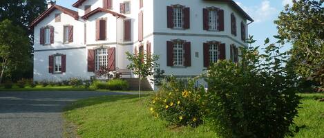 A large, two-story building with red shutters and a white facade, surrounded by lush greenery and a gravel path.