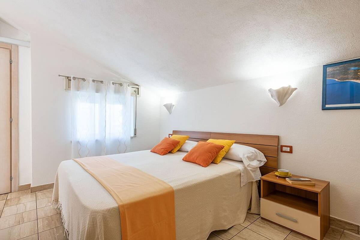 A bedroom with a bed, curtains, a nightstand, a wall-mounted lamp, and a painting.