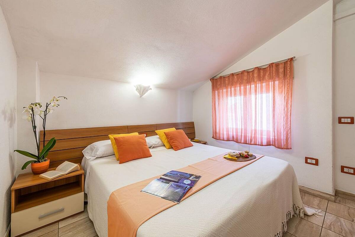 A hotel room with a bed, a nightstand with a plant, a window with curtains, and a wall-mounted light.