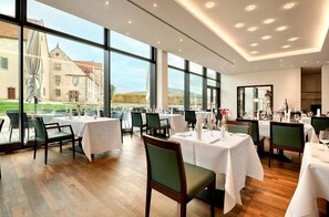 Un restaurante con mesas dispuestas para comer, amplias ventanas que ofrecen vistas a un edificio y follaje, y una decoración interior moderna.