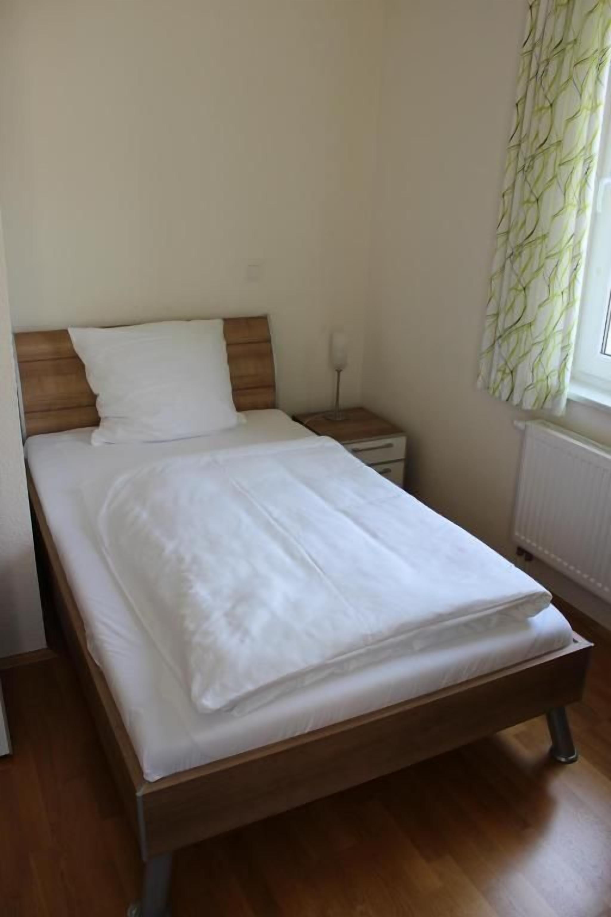 A single bed with a wooden headboard, a bedside table, and a window with a curtain.