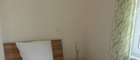 A single bed with a wooden headboard, a bedside table, and a window with a curtain.