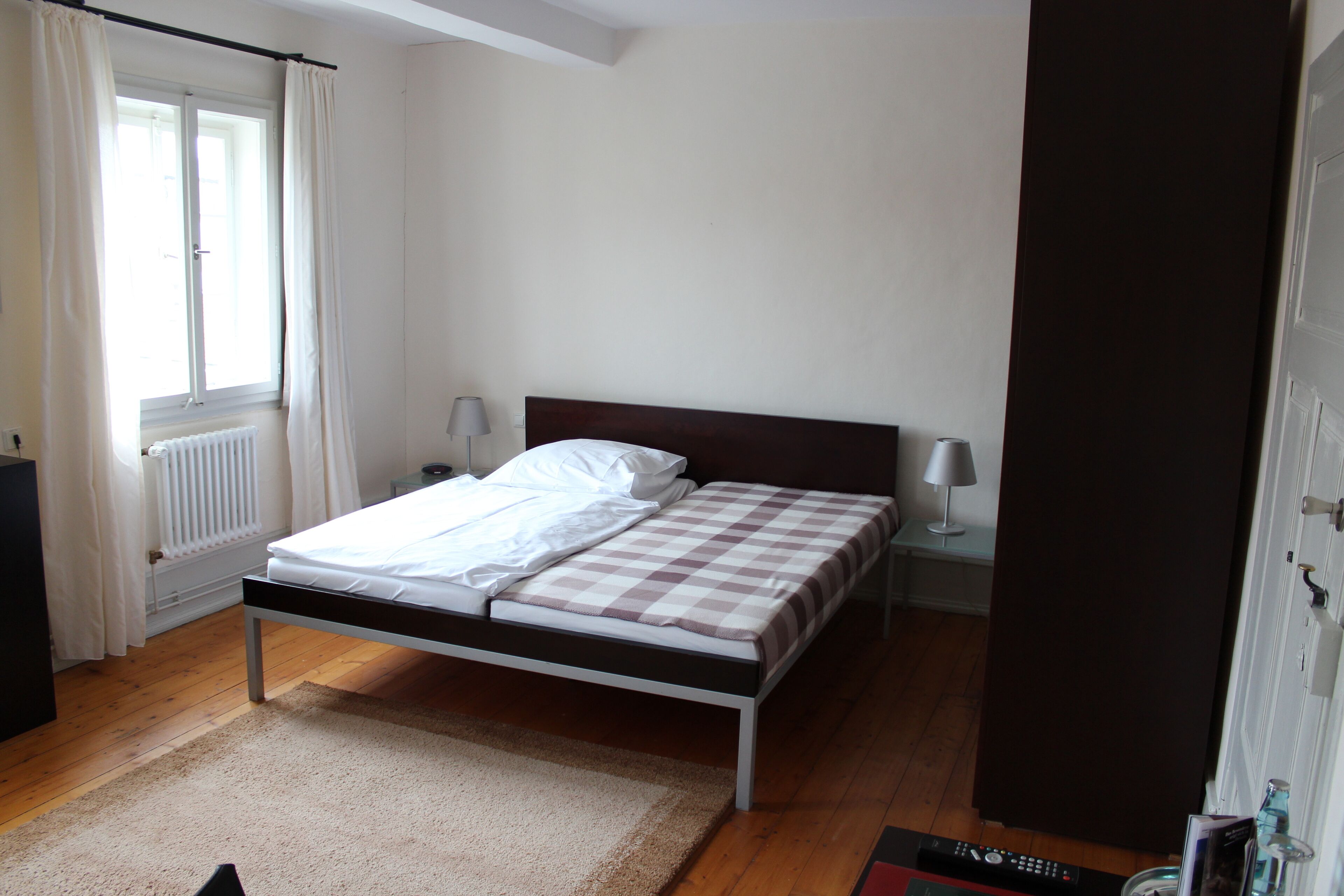 A bedroom with a bed, a radiator, a window with curtains, a bedside table with a lamp, and a door.