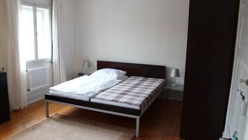 A bedroom with a bed, a radiator, a window with curtains, a bedside table with a lamp, and a door.