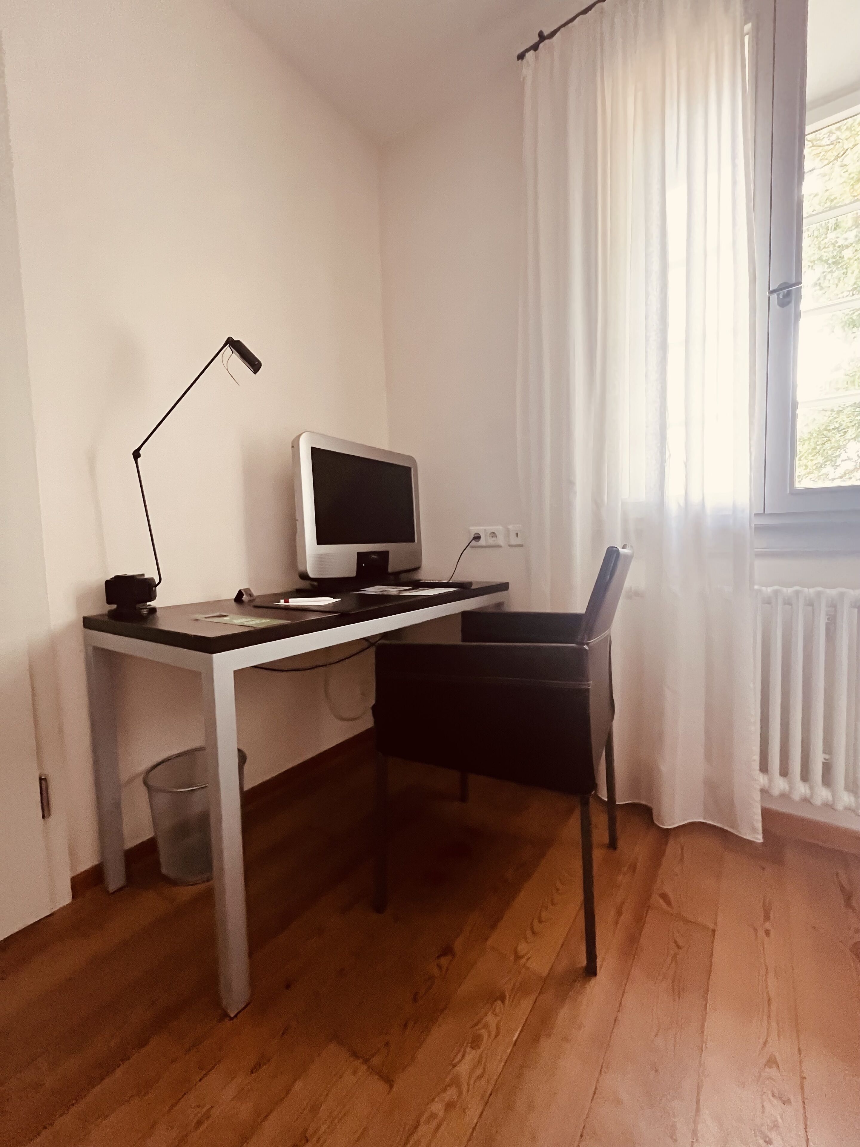 Une petite pièce au mobilier minimaliste, avec un plancher en bois, un bureau doté d’un ordinateur et d’une lampe, une chaise et une fenêtre ornée de rideaux blancs.