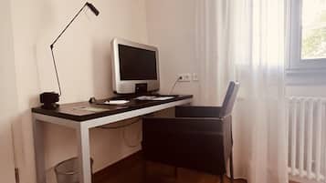 Una habitación pequeña y escasamente amoblada, con piso de madera, un escritorio con computadora y lámpara, una silla y una ventana con cortinas blancas.