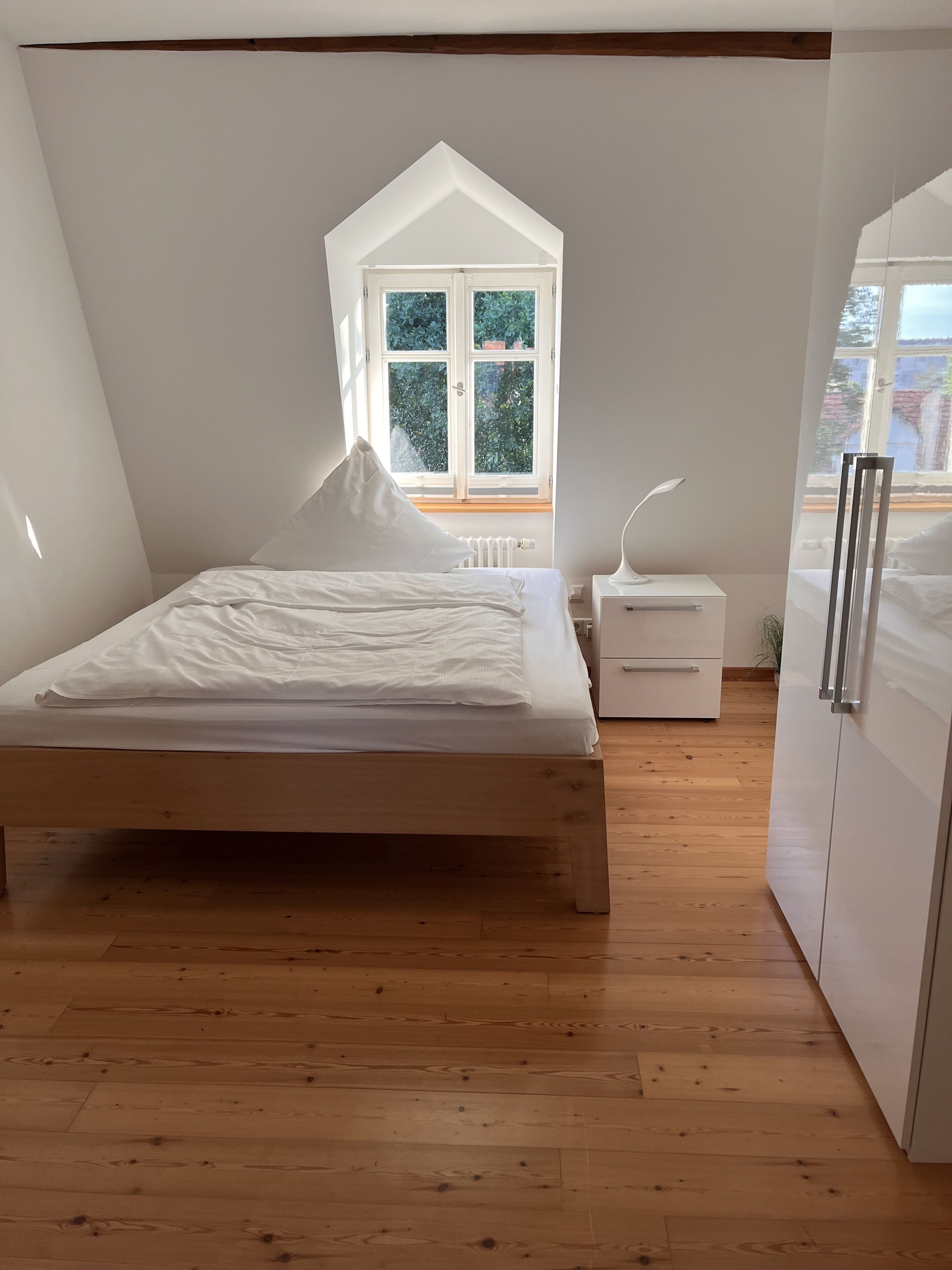 Un dormitorio con cama de madera, mesita de noche blanca, cómoda blanca y ventana con vista a follaje.