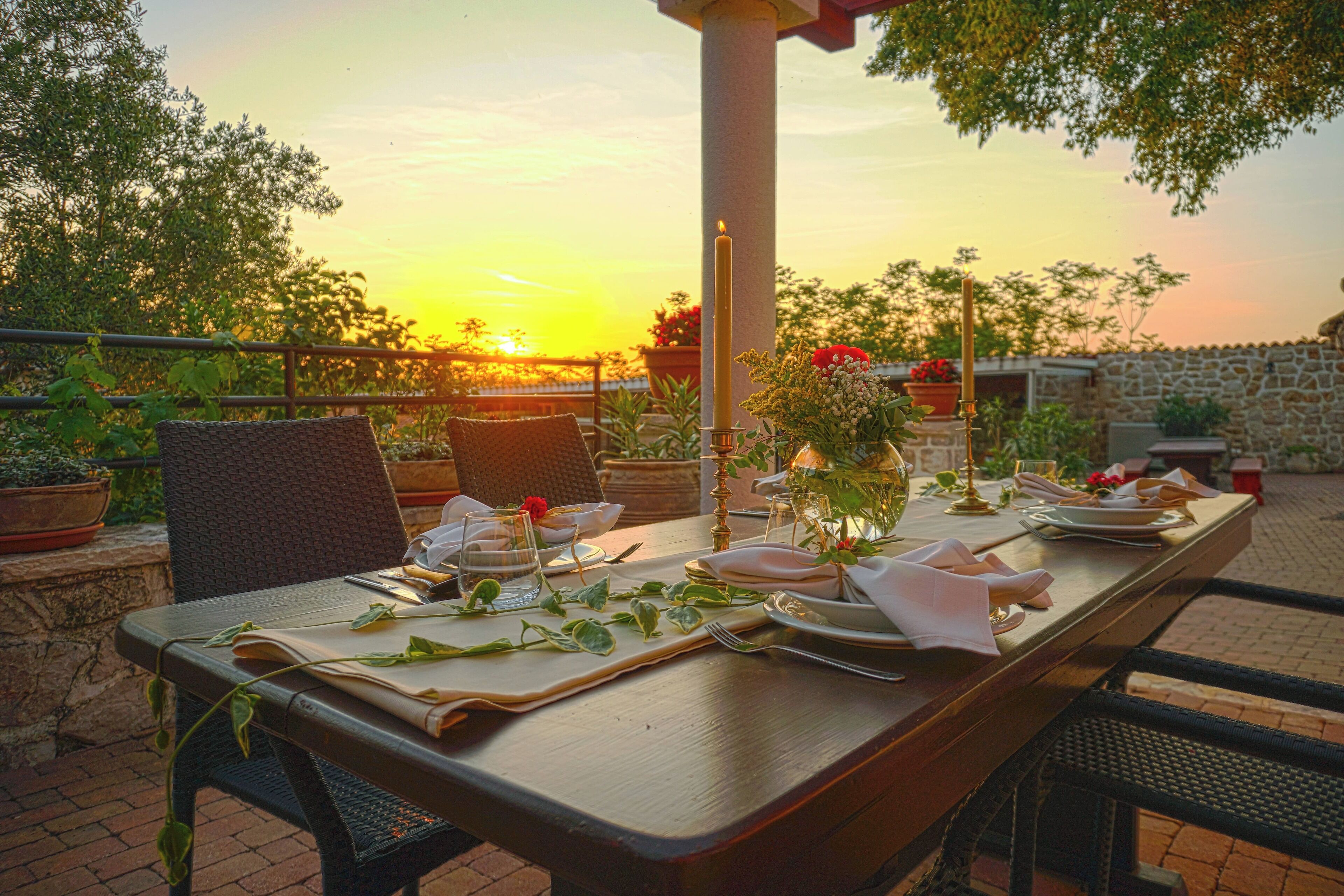 An outdoor dining table set for two with a candle centerpiece, surrounded by potted plants and a stone wall.