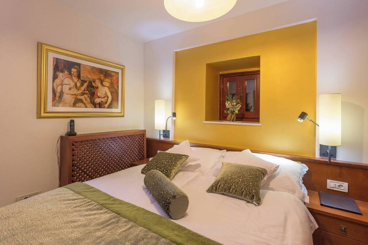 Una camera d'albergo con un letto, un quadro appeso al muro, una finestra con vista e un comodino con una lampada.