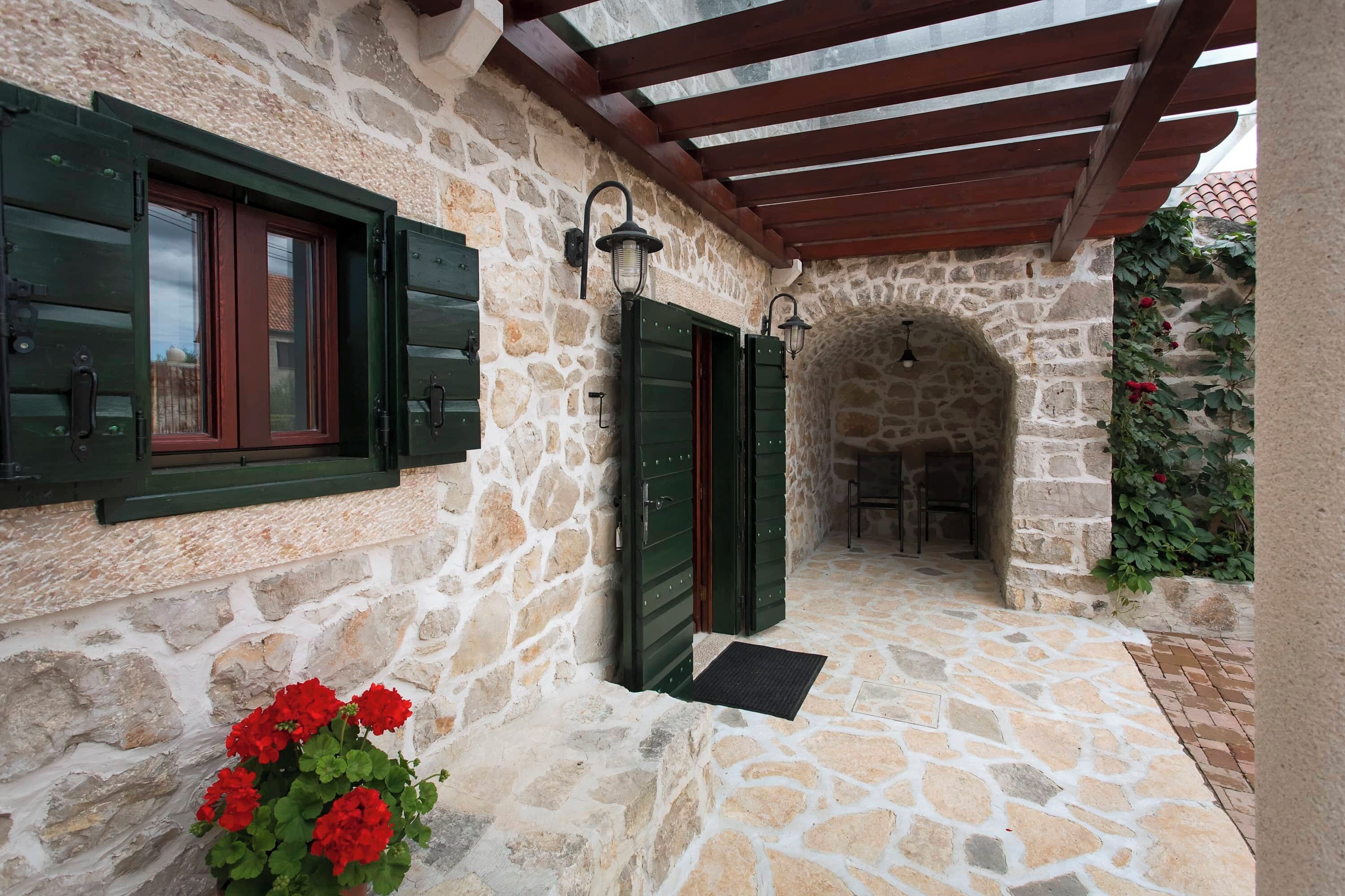 A stone building with a covered entrance, green shutters, and a potted plant with red flowers.