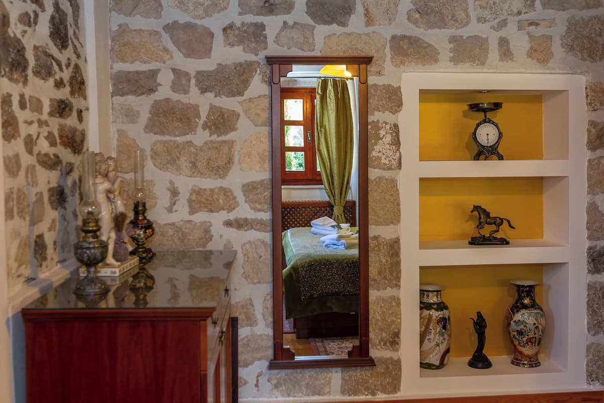 Una stanza con una parete in pietra, uno specchio che riflette un letto con copripiumino verde, uno scaffale con oggetti decorativi e un armadio in legno.