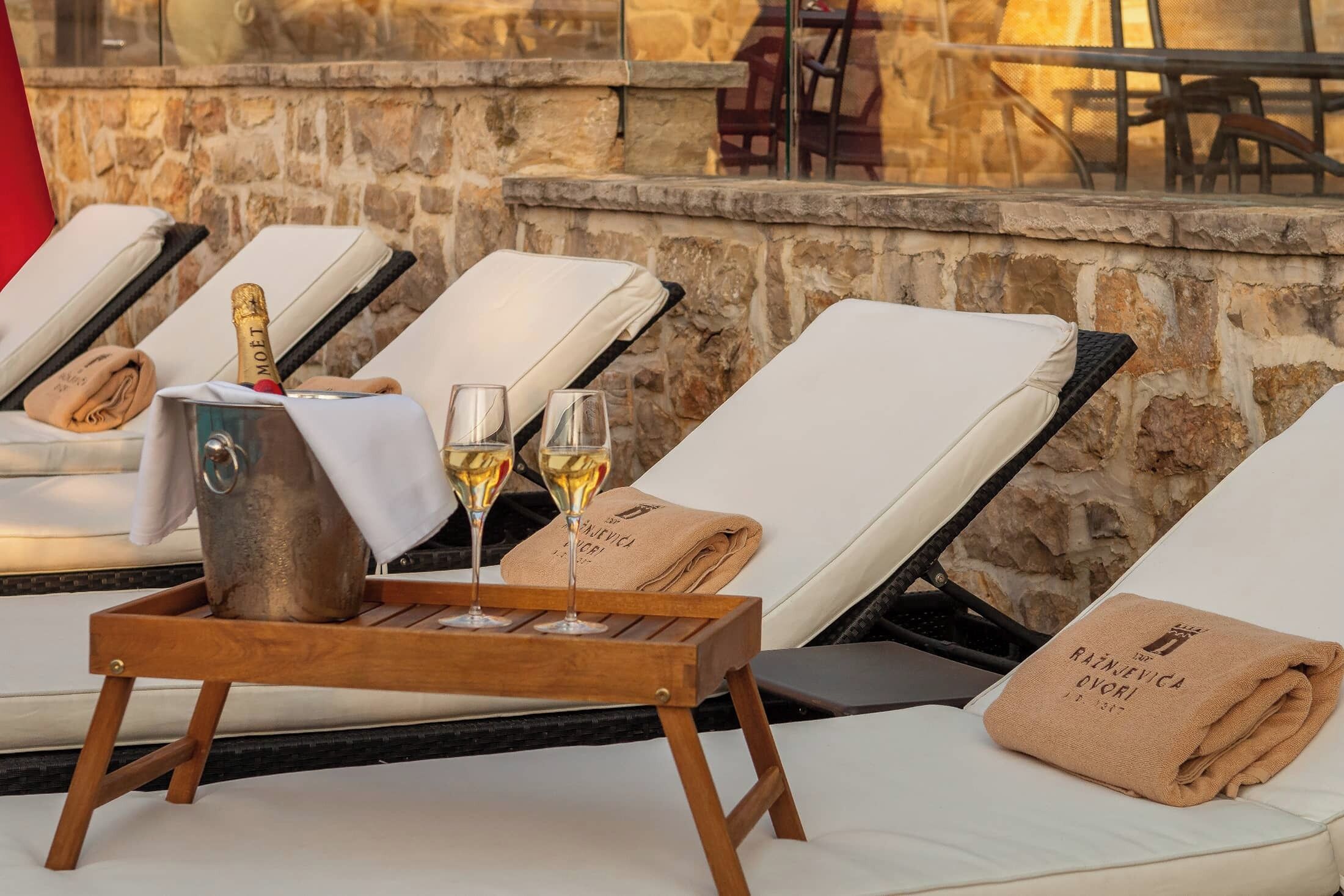 A poolside area with lounge chairs, a metal bucket with a bottle of champagne, and glasses of champagne.
