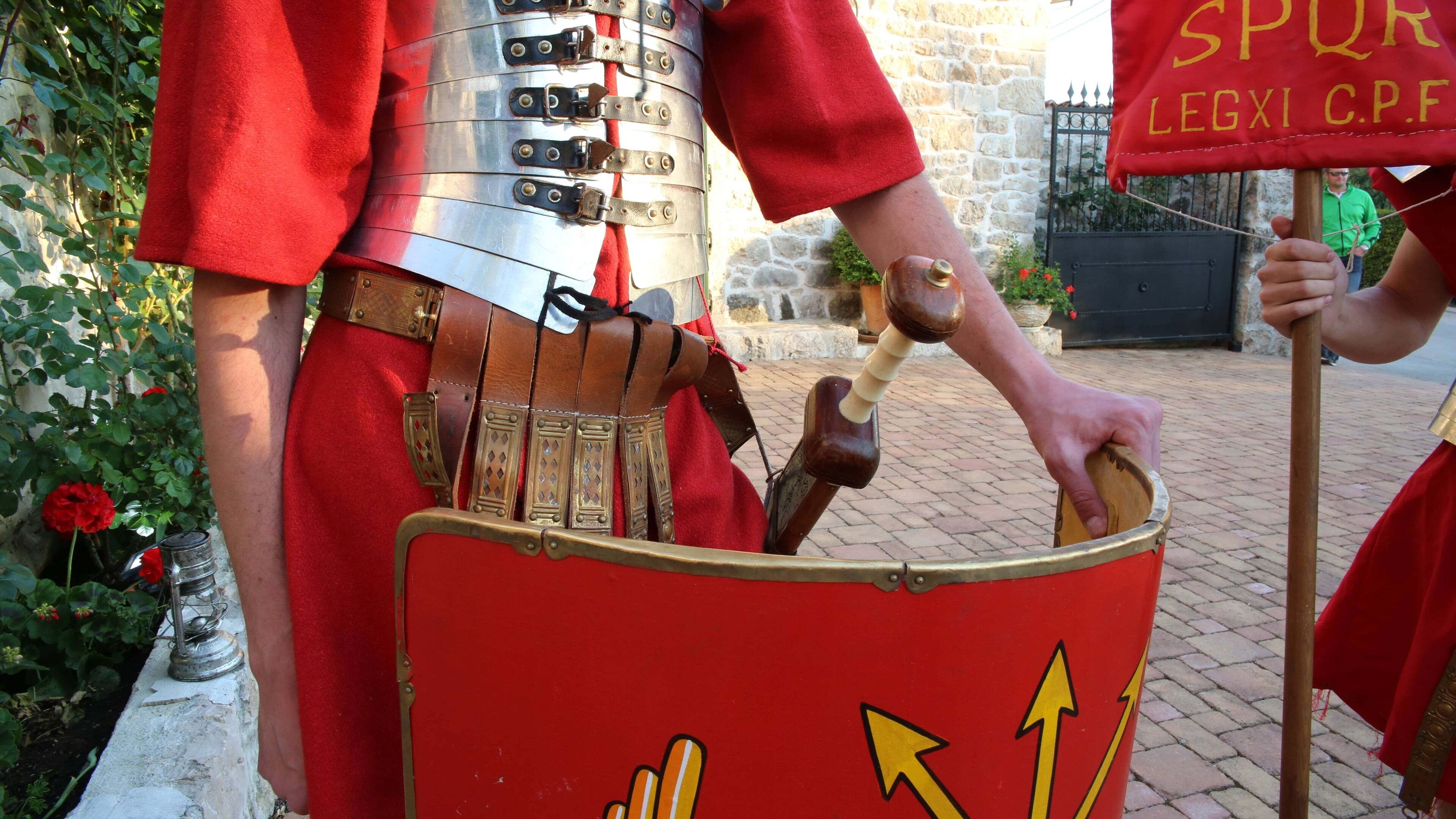 Une personne vêtue d’un costume médiéval, tenant un bouclier orné de flèches et d’un marteau.
