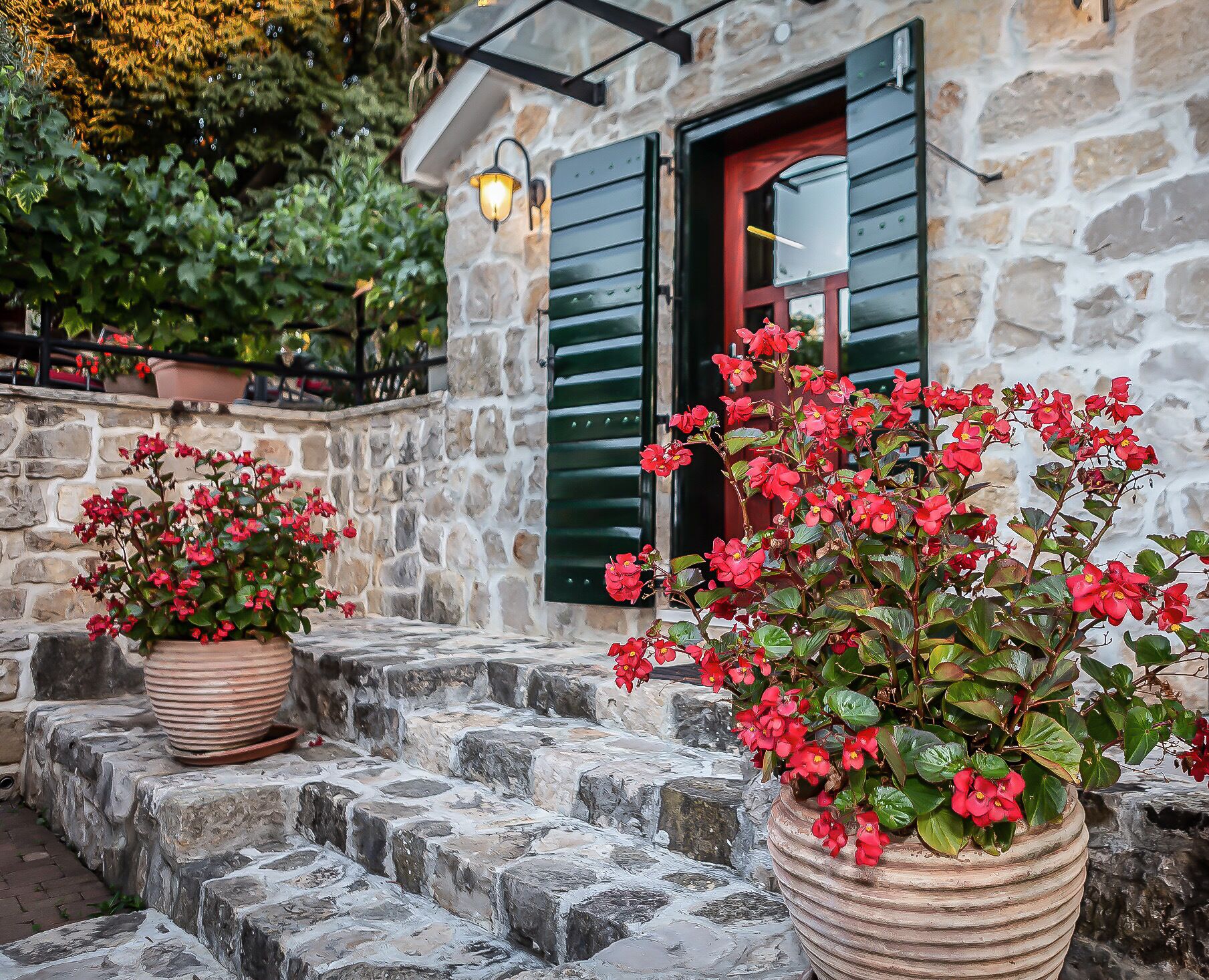 A stone building with a wooden door and green shutters. There are potted plants with red flowers in front of the entrance.
