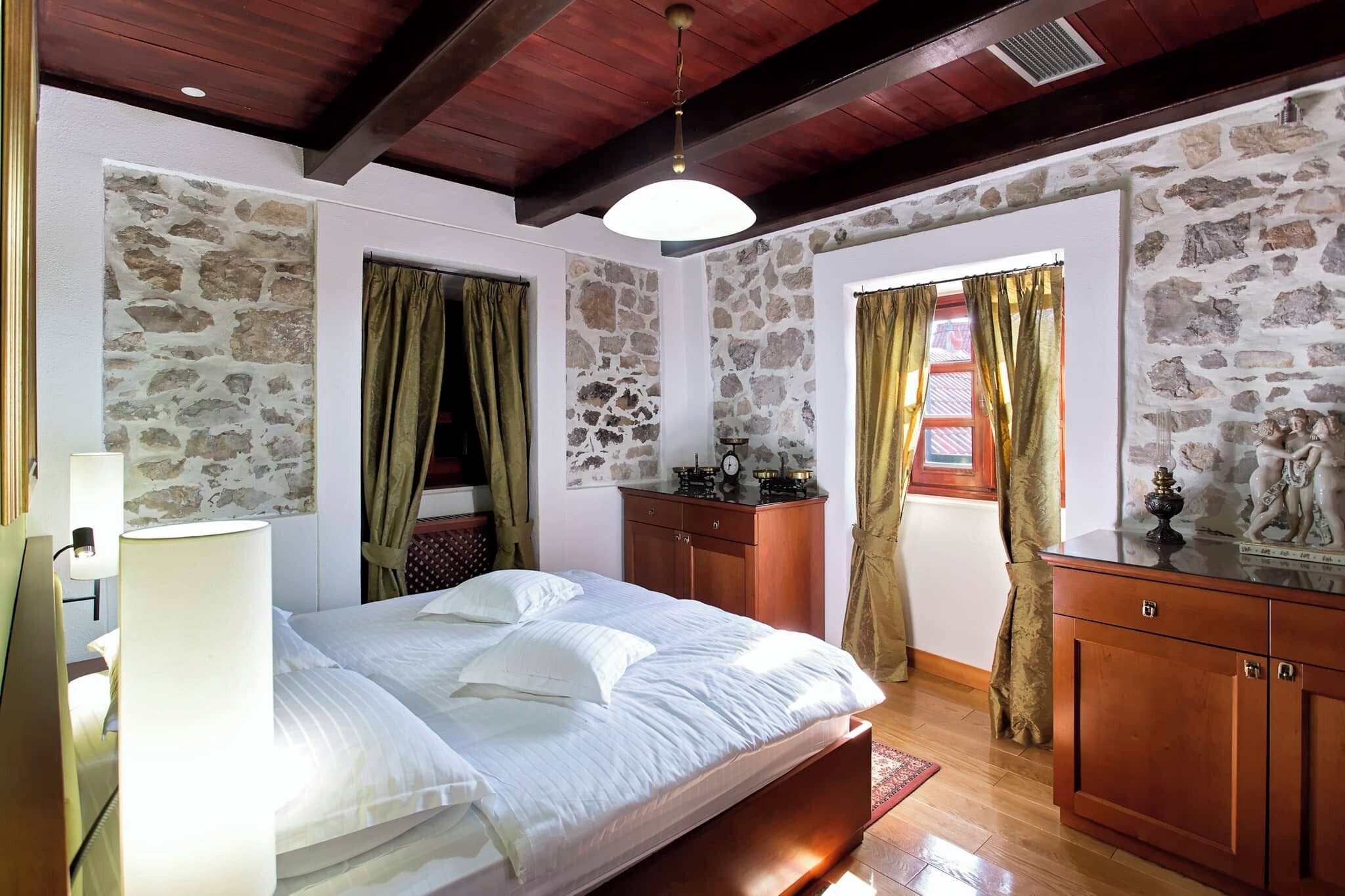 Ein Schlafzimmer mit einem Holzbett, Steinwänden, einer Holzkommode und einem Fenster mit Vorhängen.