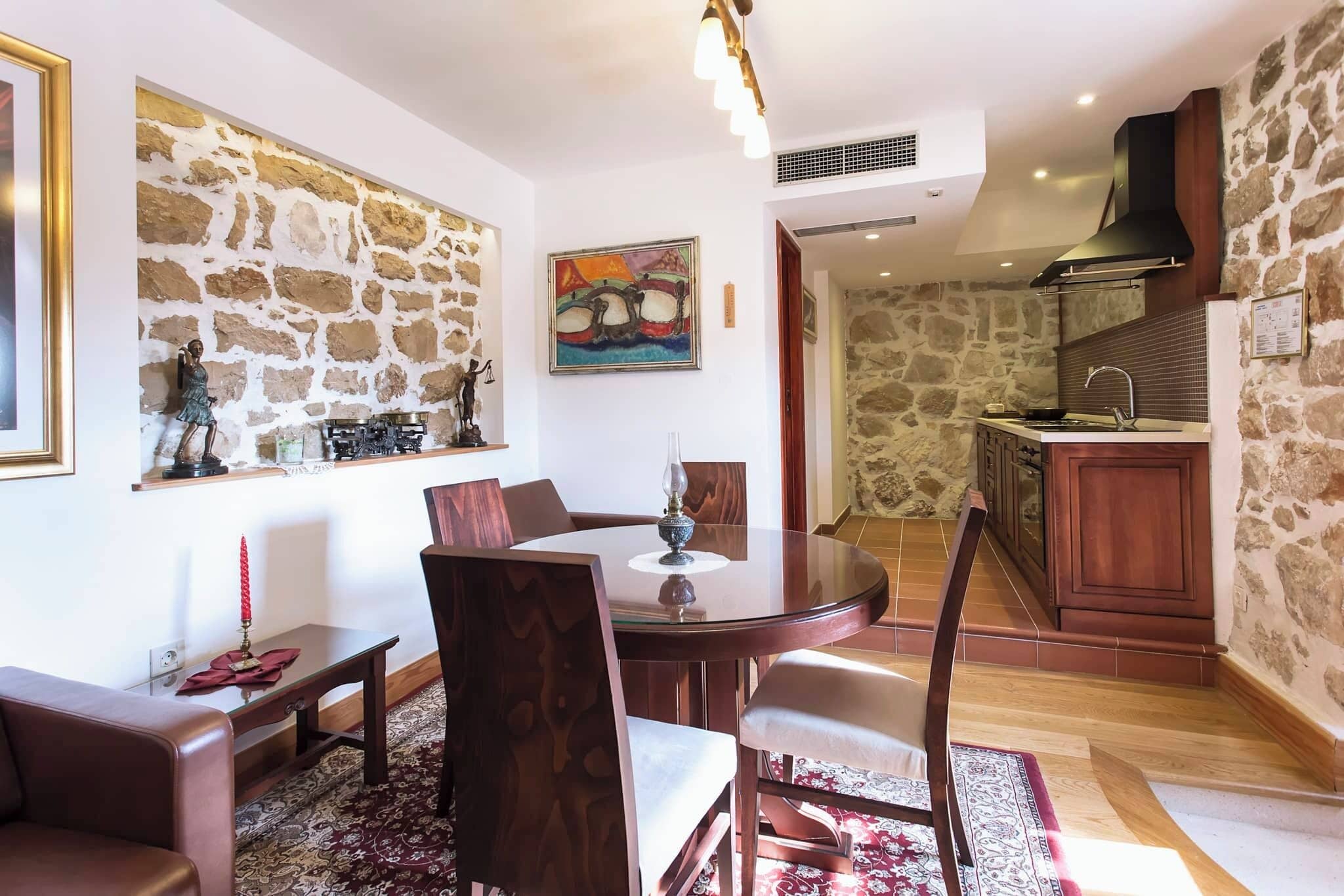Un comedor con una mesa redonda, sillas y una vela roja sobre una mesa de vidrio. Se observa una pared de piedra con un cuadro y una estatua. En el fondo, se distingue una cocina con salpicadero de piedra y armarios de madera.