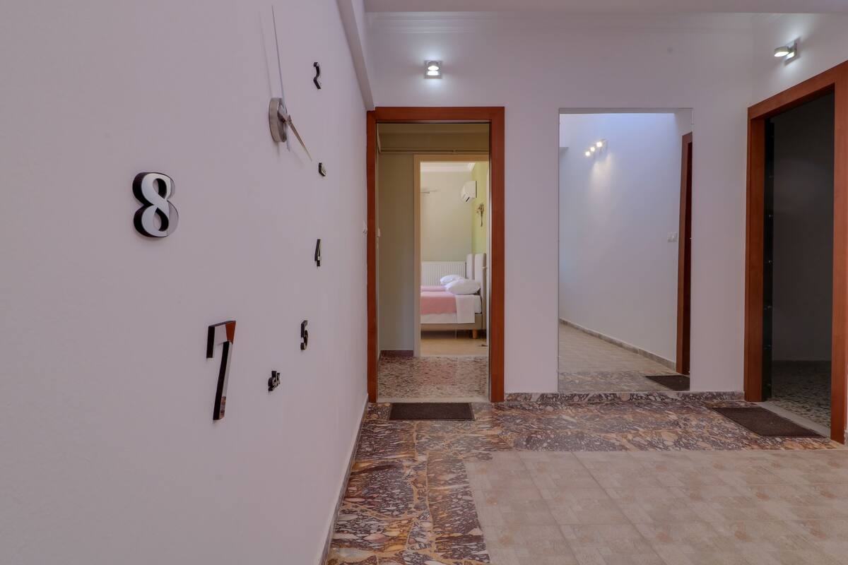 Un pasillo con puertas numeradas, suelo de baldosas y vista a un dormitorio con cama y radiador.