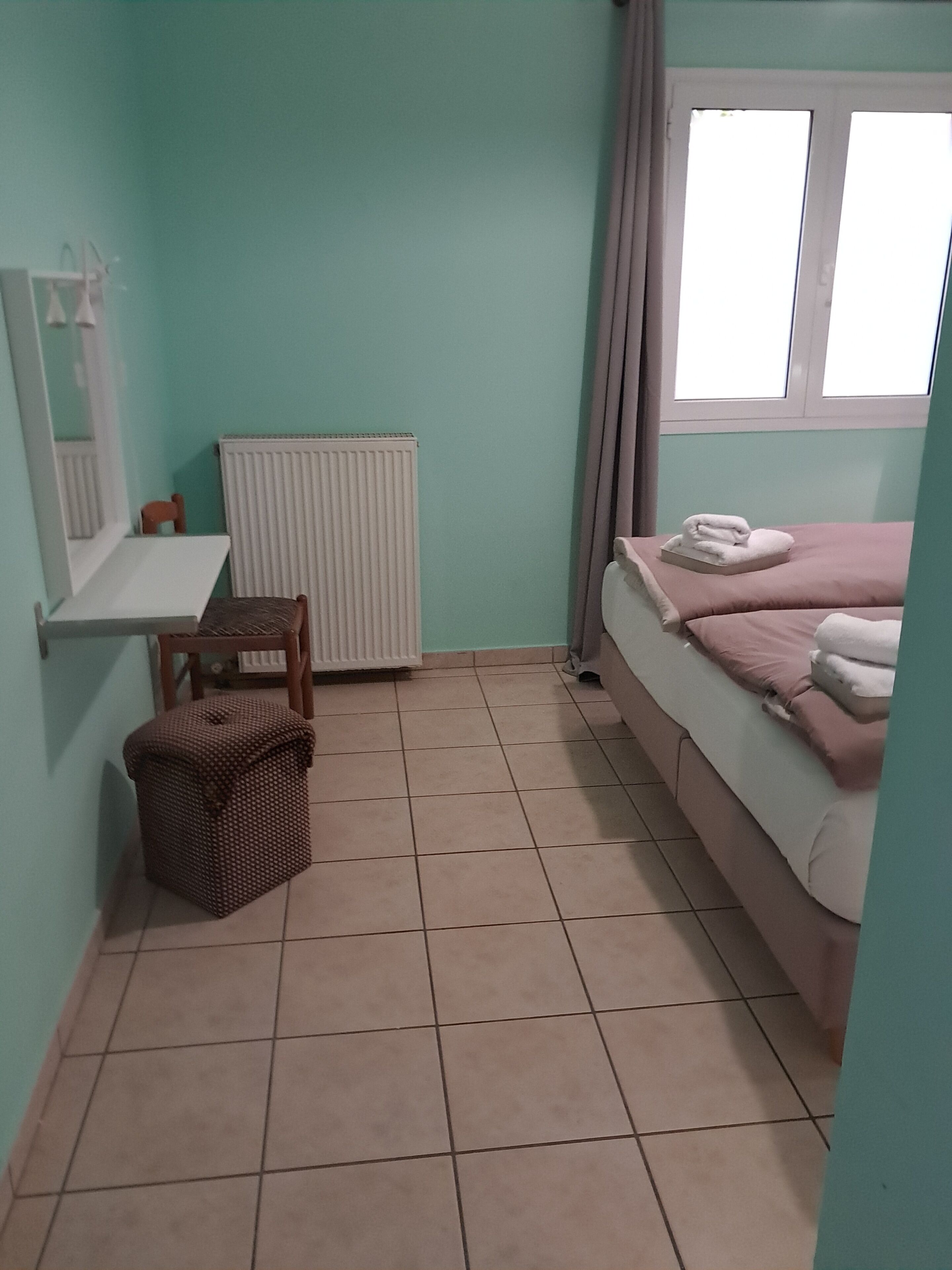 Una habitación pequeña y ordenada con una cama, un radiador, una ventana con cortinas, una mesita y una silla.