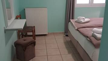 Una habitación pequeña y ordenada con una cama, un radiador, una ventana con cortinas, una mesita y una silla.