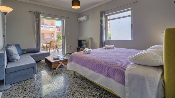 A modern hotel room with a bed, sofa, TV, and a balcony with a table and chairs. - Carpe Diem Apartments By Athens Airport (Markopoulo Mesogaias)