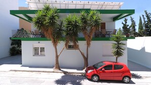 A modern two-story building with a balcony, a red car parked in front, and palm trees. - Carpe Diem Apartments By Athens Airport (Markopoulo Mesogaias)