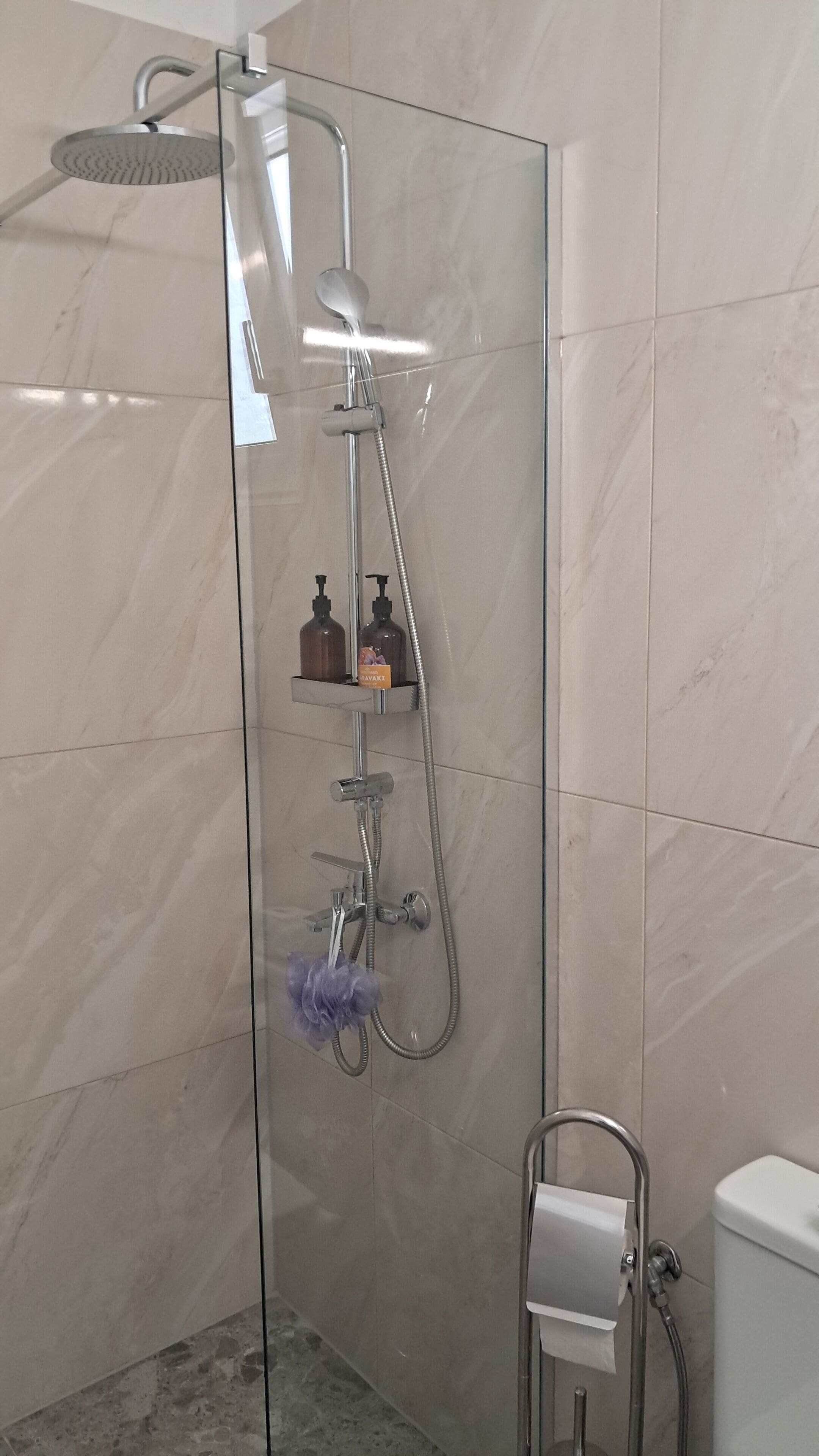 A glass-enclosed shower with a handheld showerhead, a handheld shower attachment, and a handheld shower nozzle. There is a soap dispenser and a bottle of shampoo on a shelf.