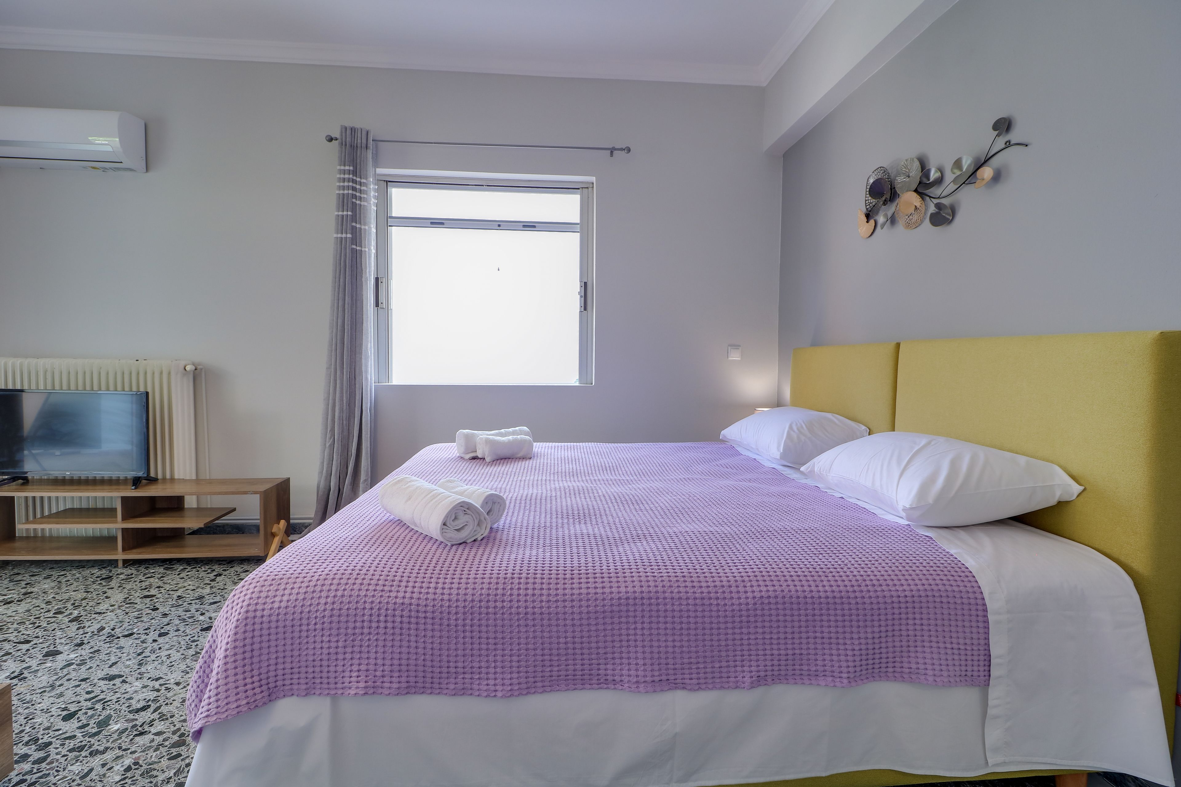 A neatly made bed with a purple checkered bedspread and white pillows, a television on a wooden stand, and a window with a curtain.