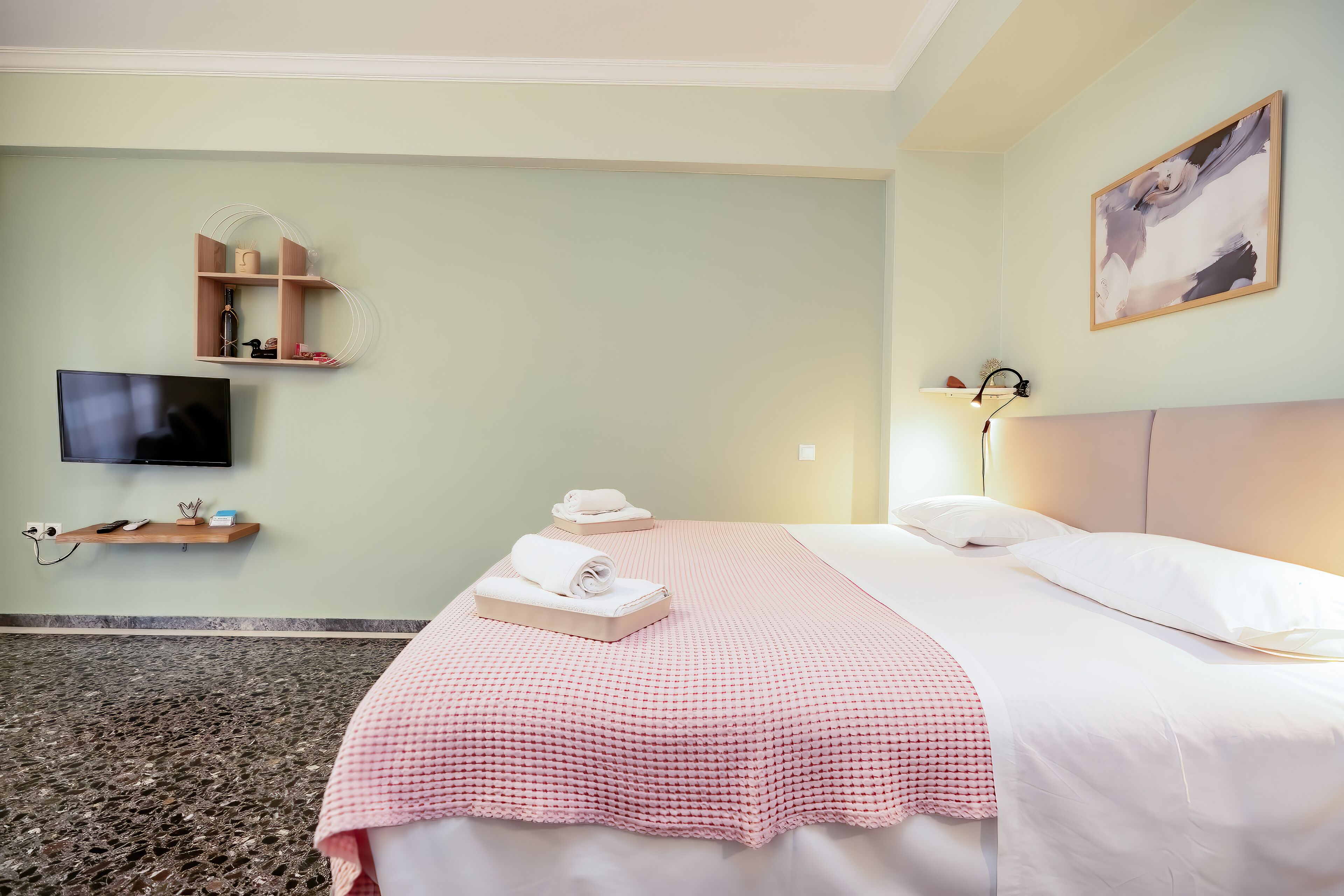 Une chambre d’hôtel avec un lit, une télévision, une étagère fixée au mur où sont rangés les articles de toilette, et un tableau.