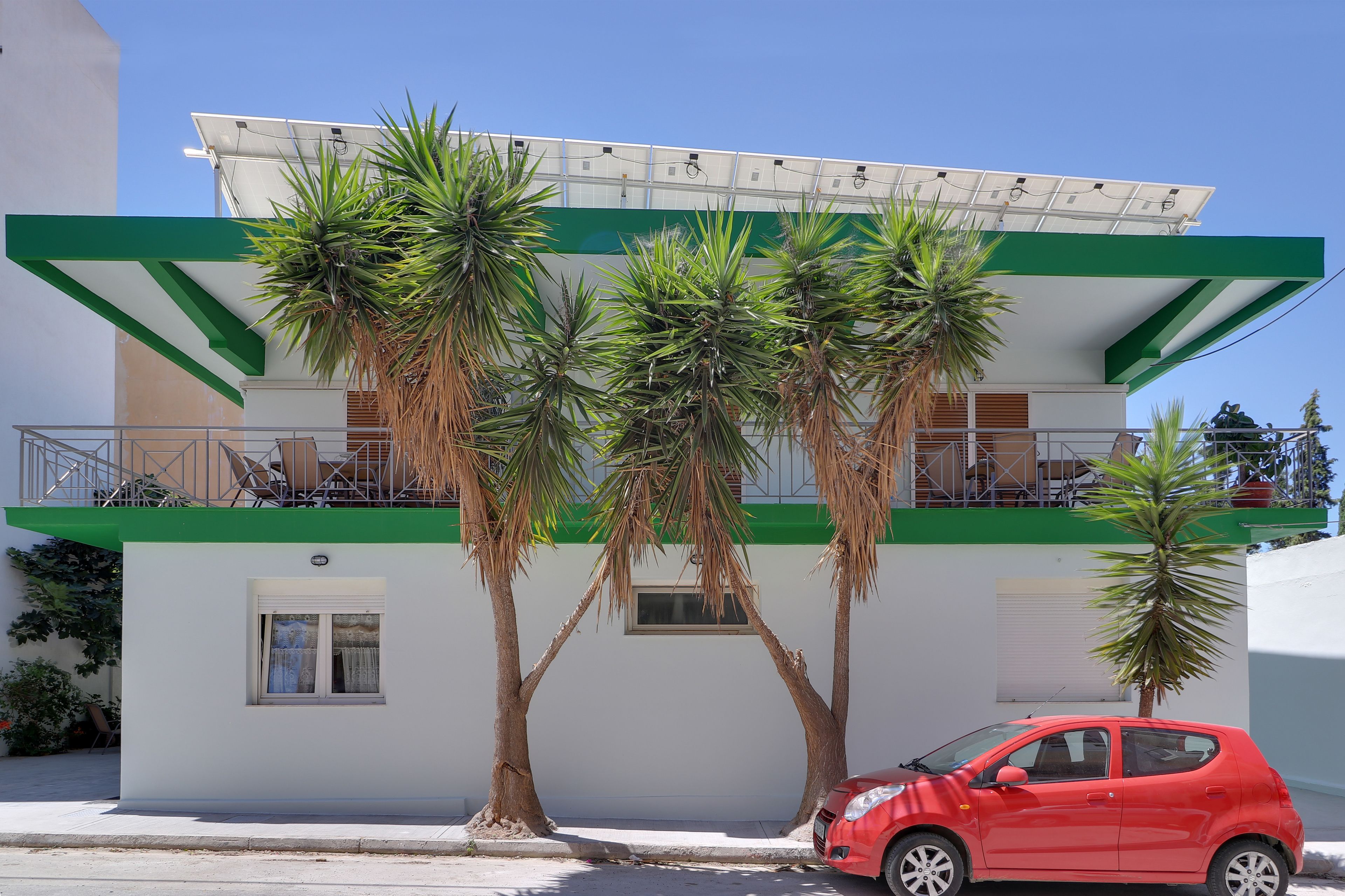 A modern building with a green and white facade, a balcony with a solar panel, and a red car parked in front.
