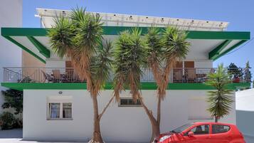 A modern building with a green and white facade, a balcony with a solar panel, and a red car parked in front.