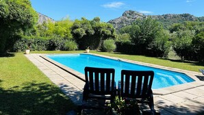 A rectangular swimming pool with two black lounge chairs facing it, surrounded by well-maintained grass and trees, with a mountainous backdrop. - Gîte La Ferme de Cabanis de Labahou SPA 8 pers (Anduze)