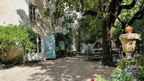 A courtyard with a gravel path, a rustic building with a blue door, and a large tree. - Gîte La Ferme de Cabanis de Labahou SPA 8 pers (Anduze)