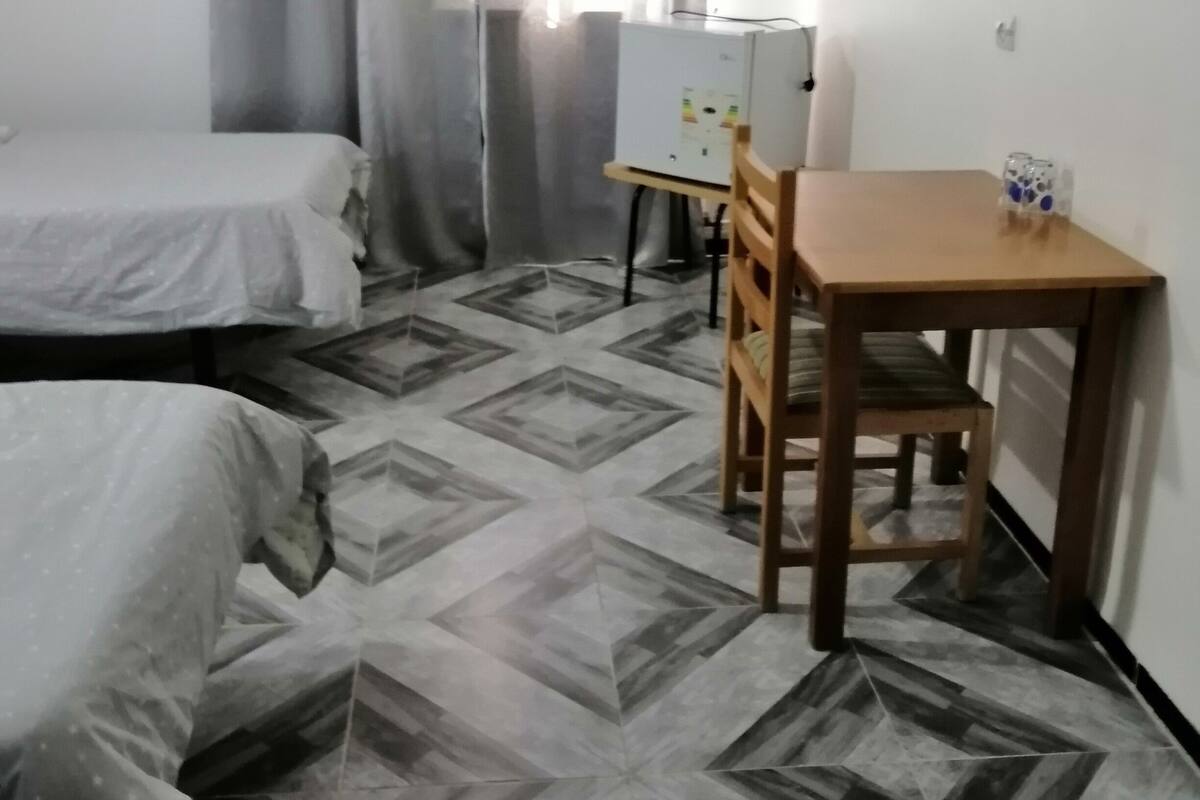 A small, clean room with two beds, a wooden table with a chair, a TV mounted on the wall, and an air conditioning unit.