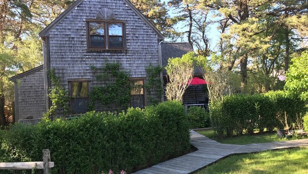 A wooden house with a gabled roof, surrounded by dense greenery and a wooden pathway leading to the entrance. - 2 Bed/2 Bath on 1/4 acre w/fenced yard, short walk to 40th thru rolling dunes. (Nantucket)