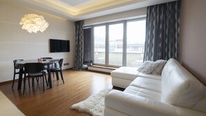 Una sala de estar moderna con un sofá blanco, una mesa de comedor y sillas. Hay un ventanal con cortinas, un televisor de pantalla plana montado en la pared y una alfombra mullida en el suelo.