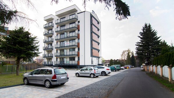 A multi-story residential building with balconies, parked cars along the street, and a clear sky.
