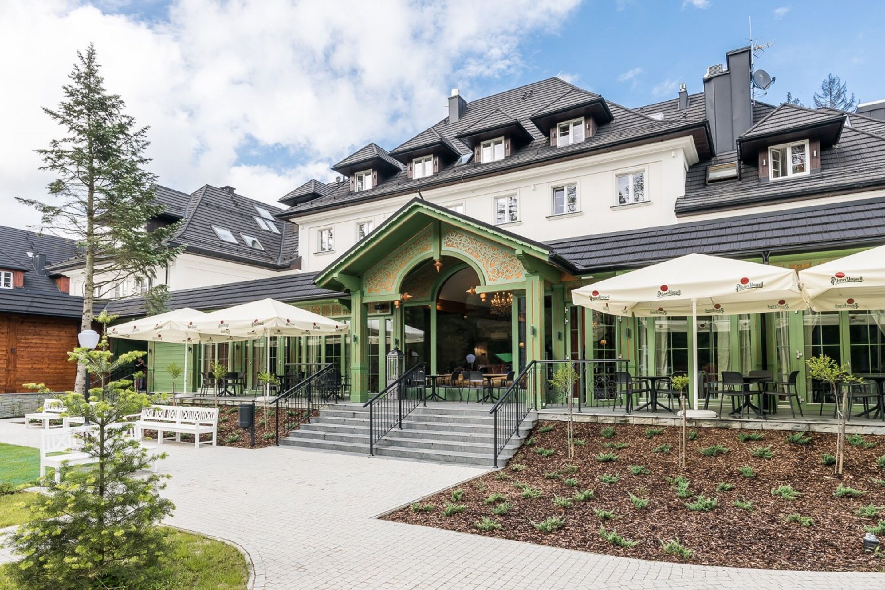 A building with a green facade, outdoor seating area with umbrellas, and a staircase leading to the entrance.