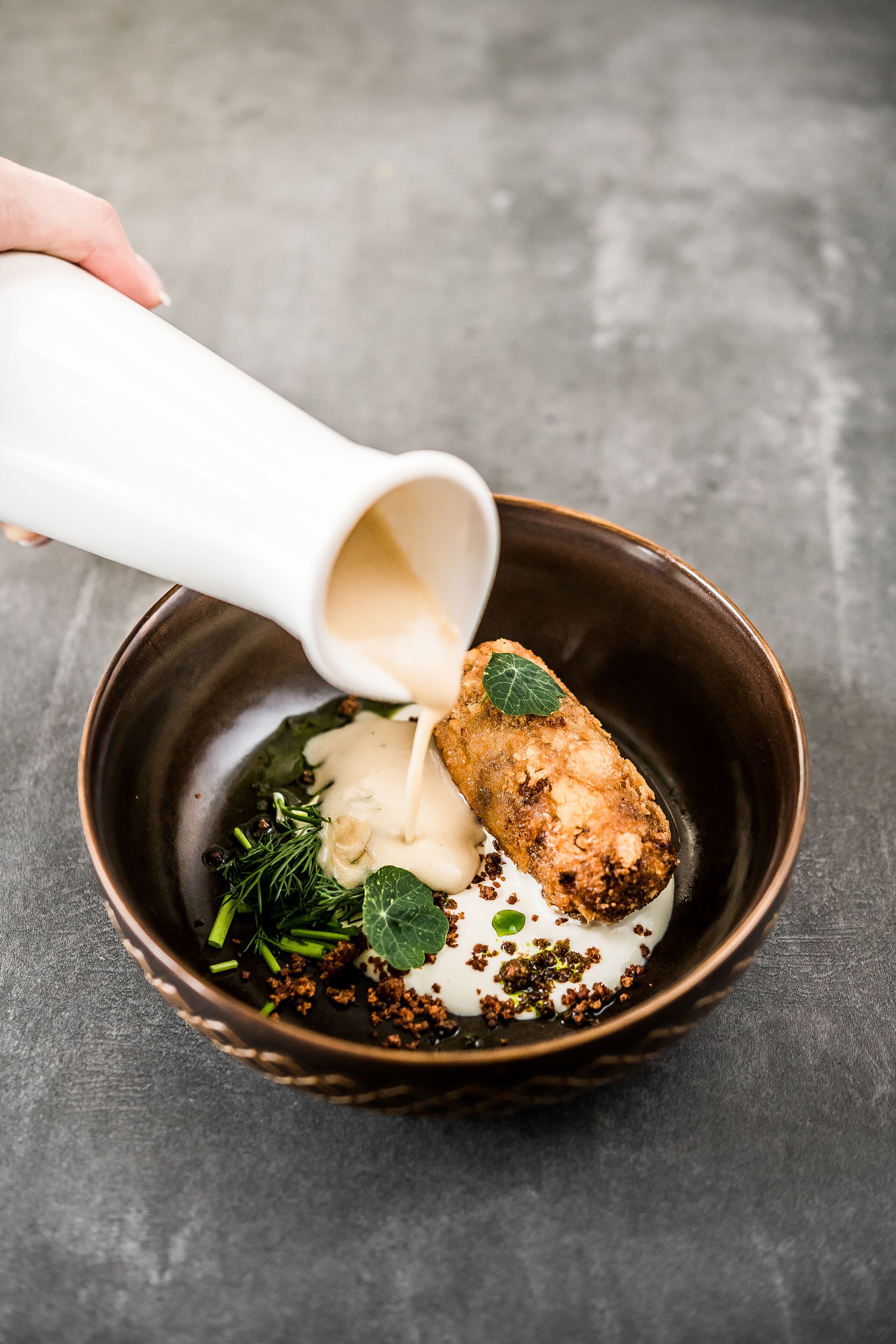 A hand pouring a creamy sauce over a dish with a fried item, garnished with herbs and spices.