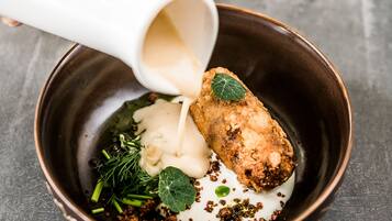 A hand pouring a creamy sauce over a dish with a fried item, garnished with herbs and spices.