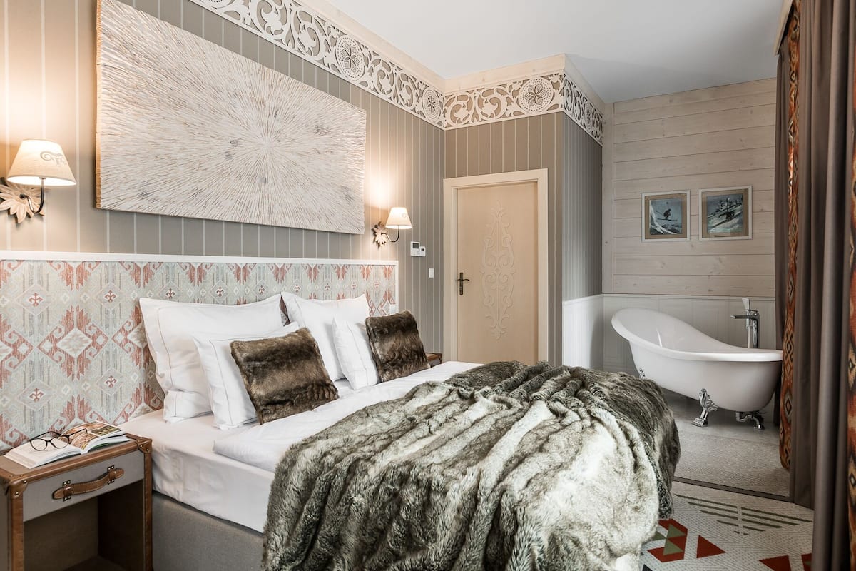 Un dormitorio con una cama grande, una mesita de noche, una lámpara de pared, un cabecero decorativo, un espejo grande y una bañera.