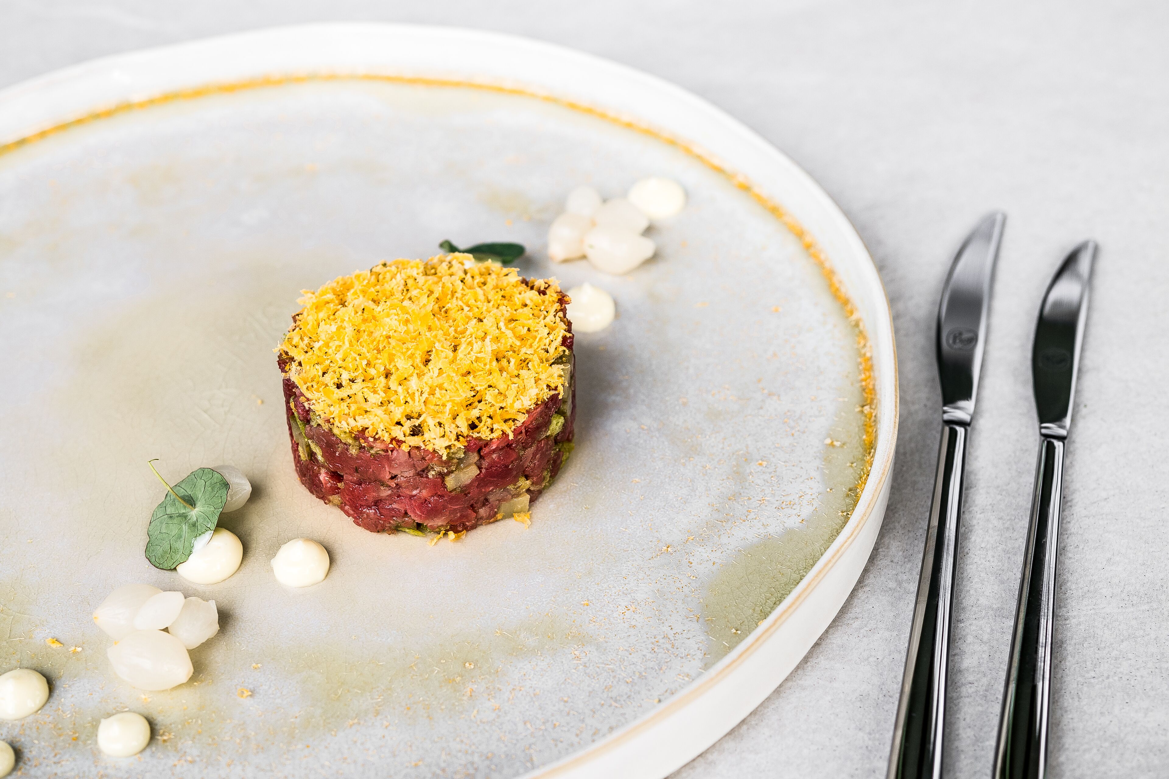 A plated dish with a golden-brown crust, garnished with white spherical elements and a green leaf, on a white plate with a subtle rim.
