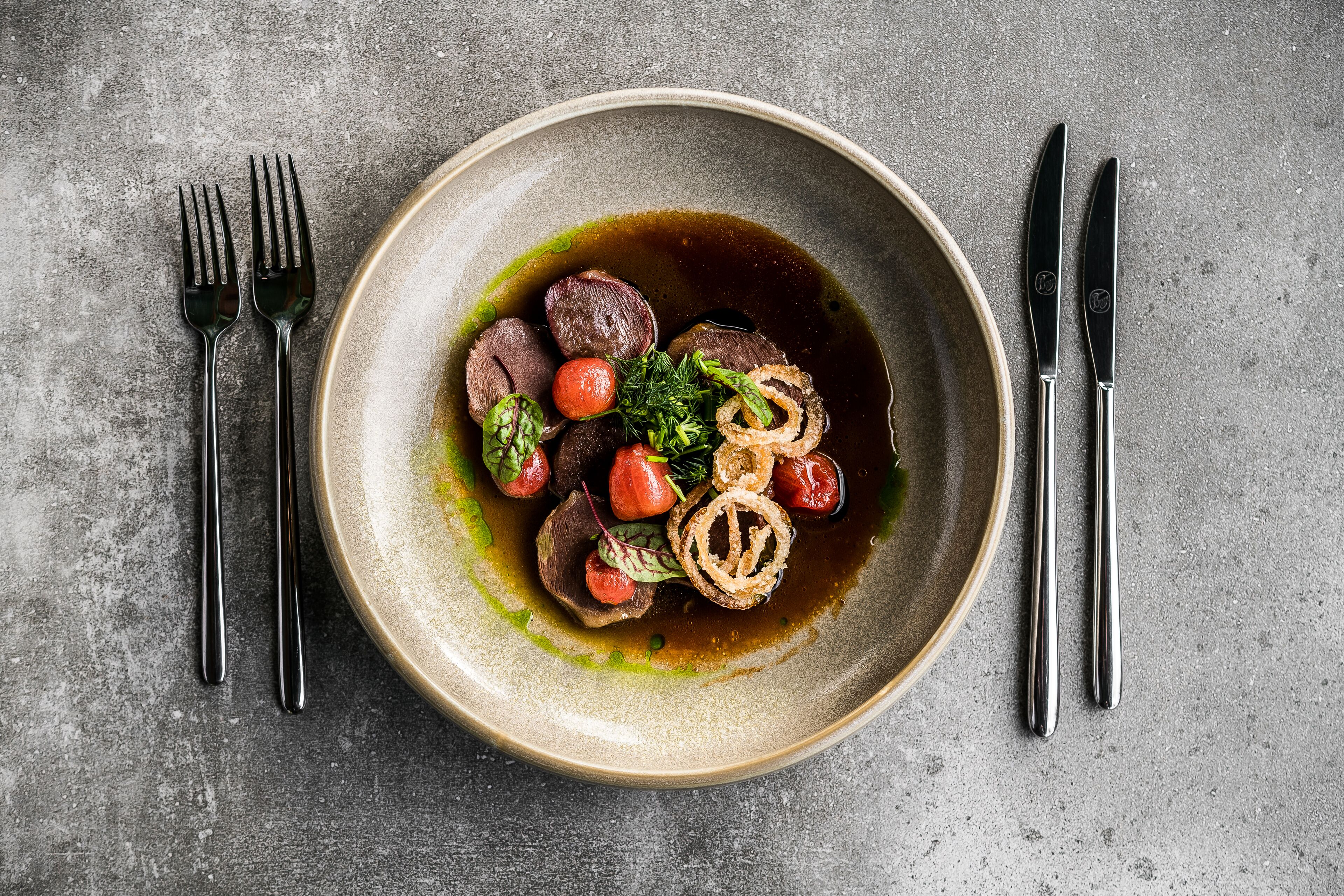 A bowl of food with a dark sauce, garnished with herbs and vegetables, placed on a textured surface with cutlery beside it.