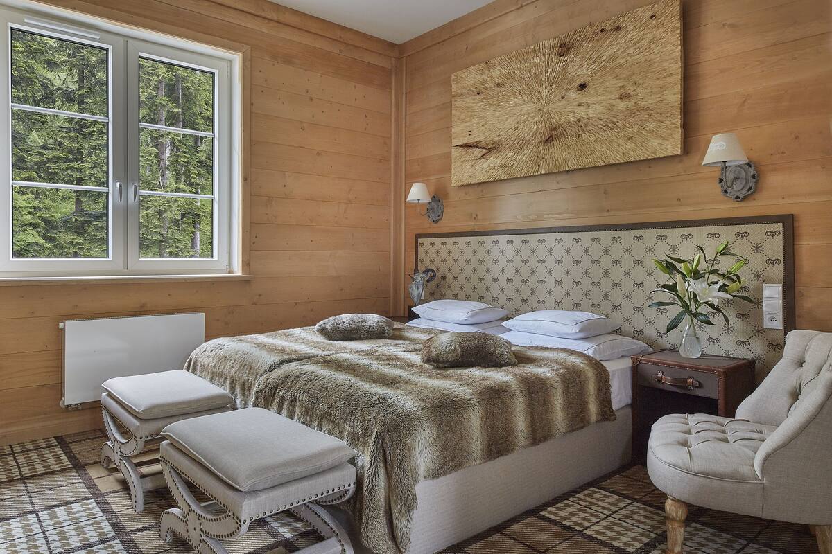 Una habitación con paneles de madera, una cama, una silla, una mesita de noche y un ventanal con vistas a árboles.