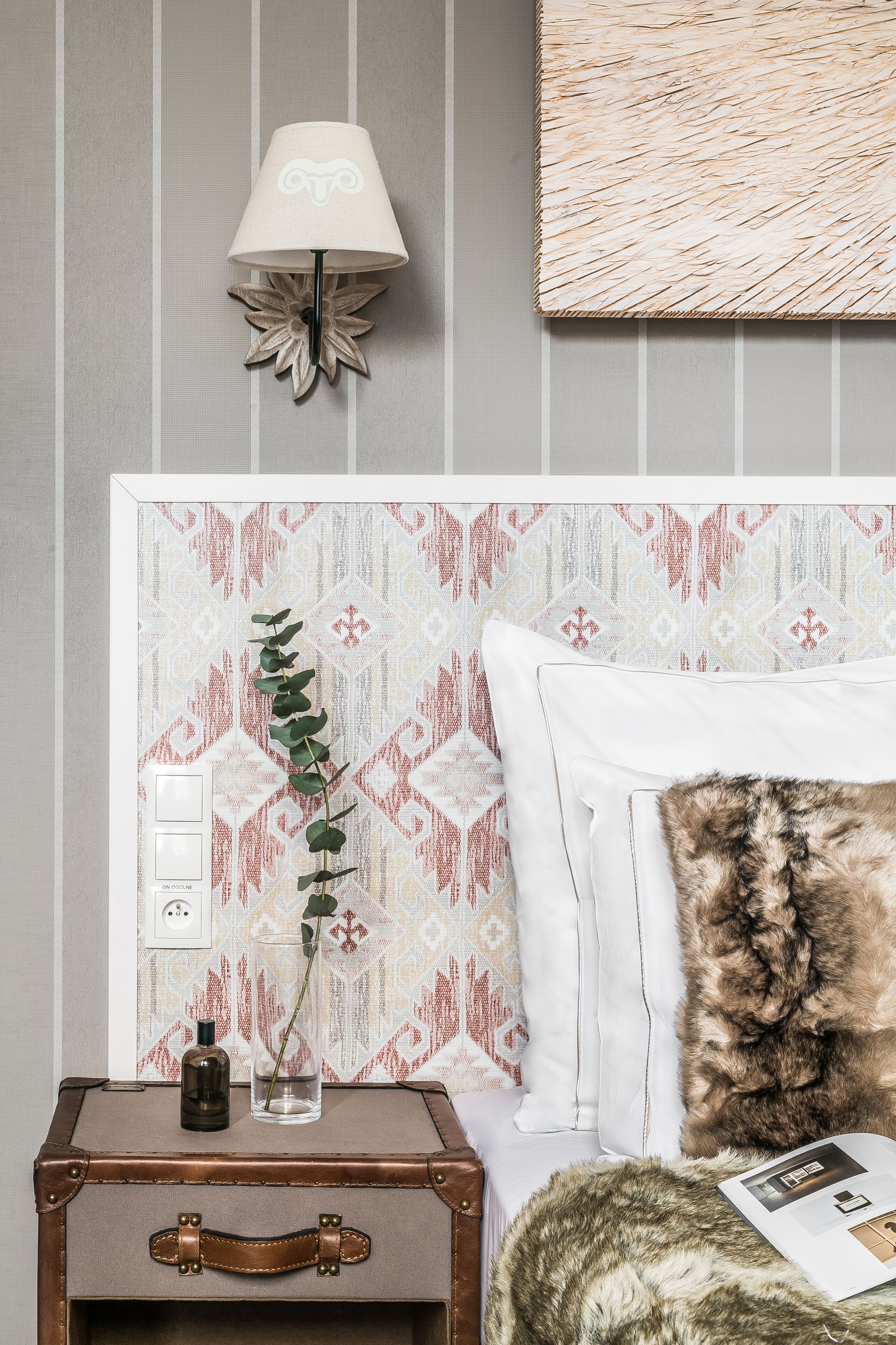 A neatly arranged bedroom with a patterned headboard, a bedside lamp, a small table with a bottle and glass, and a suitcase.