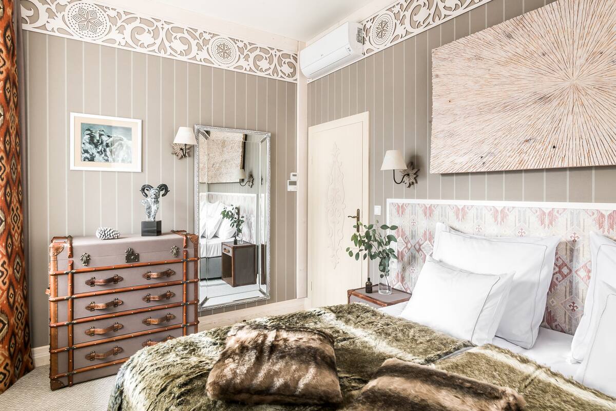 Un dormitorio con papel tapiz a rayas, un espejo grande, un aparador de madera, una cama con ropa blanca y un adorno en la pared.